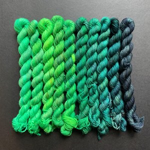 May include: A collection of eight skeins of yarn in varying shades of green, from bright lime to deep teal. The yarn is tightly twisted, and the skeins are arranged in a row, showcasing a gradient of color.