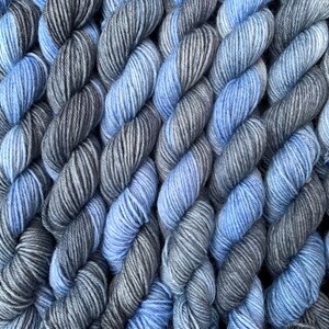 May include: Close-up of a collection of yarn skeins in shades of blue and gray. The yarn is tightly wound, creating a textured surface. The color gradient ranges from light blue to dark gray.