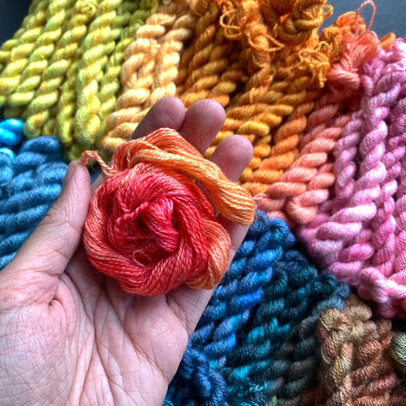 May include: A hand holding a bundle of orange and red embroidery floss, with a variety of colorful floss bundles in the background. The floss is arranged in a gradient of colors, including yellow, blue, pink, and orange.