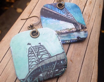 May include: A pair of square-shaped earrings with a watercolor image of a bridge. The earrings are made of metal and have a bronze-colored hook. The image on the earrings is a light blue and green watercolor painting of a bridge.