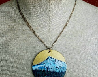 May include: A gold pendant necklace with a round, painted pendant featuring a blue mountain scene with a lake and trees. The pendant is on a gold chain.
