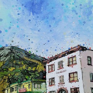 May include: A painting of two buildings with a mountain in the background. The building on the right is white with red trim and says "Van Gilder Hotel". The building on the left is yellow with green trim and says "Liberty Theater". The sky is blue with white and blue splatter paint.