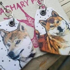 May include: A pair of earrings featuring a white ceramic tag with a black and white illustration of a Shiba Inu dog. The other earring features a white ceramic tag with a brown and white illustration of a Shiba Inu dog.