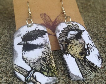 May include: A pair of dangle earrings featuring a black and white bird illustration on each earring. The earrings are made of metal and have a silver finish.