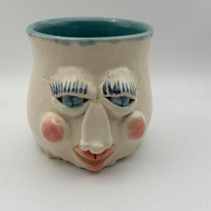Könnte beinhalten: Eine handgefertigte Keramiktasse mit einem skurrilen Gesichtsdesign. Die Tasse hat eine cremefarbene Basis mit blauen Akzenten an Rand, Augen und Wimpern. Rosa Rouge und ein roter Mund vervollständigen das Gesicht. Ein einzigartiges und künstlerisches Stück.