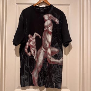 May include: A black short-sleeved t-shirt featuring a large graphic print of two figures in silver and red suits running through a dark city landscape. The shirt has a crew neckline and the text LOT29 printed on the inner collar label area.