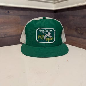 Puede incluir: Una gorra de camionero verde y blanca con una malla blanca en la parte posterior. La gorra tiene un parche en la parte delantera que dice "Remington First in the Field" con una imagen de un pato volando.