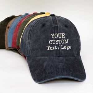 Pode incluir: Uma fileira de bonés de beisebol em várias cores, incluindo azul marinho, castanho, vermelho e amarelo. O boné azul marinho na frente tem o texto "YOUR CUSTOM Text / Logo" bordado em branco. Os bonés são feitos de um material macio e desgastado.
