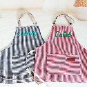 May include: Two striped aprons, one navy blue and the other red, with matching pockets and tan straps. The navy apron has "Aubrey" embroidered in teal, and the red apron has "Caleb" embroidered in green.