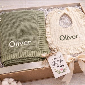 Può includere: Un set regalo per neonati in una scatola di cartone, che include una coperta lavorata a maglia verde salvia e un bavaglino color crema con bordi a balze. Entrambi gli articoli sono ricamati con il nome "Oliver". Un'etichetta regalo recita "A Special Gift for Baby".