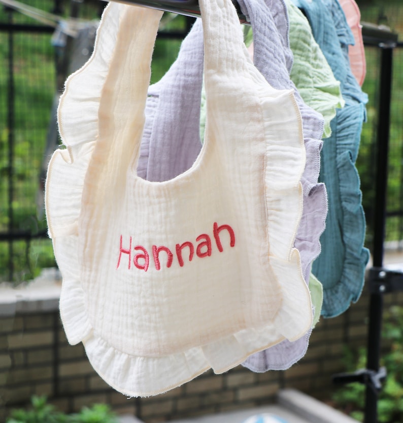以下が含まれることがあります： フリル付きの白いコットン製のベビーよだれかけ。赤い糸で「Hannah」と刺繍されています。