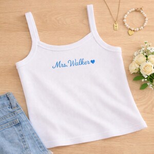 Könnte beinhalten: Weißes Camisole-Top mit dünnen Trägern. Der Text "Mrs. Walker" ist in blauer Schrift gestickt, gefolgt von einem blauen Herz. Eine Jeansshorts ist unten links zu sehen. Eine goldene Halskette und ein Perlenarmband sind im Hintergrund.