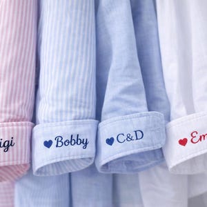 Op de afbeelding: Close-up van vier overhemden met knopen en gepersonaliseerde manchetten. De shirts zijn roze en wit gestreept, lichtblauw gestreept, lichtblauw en wit. Elke manchet heeft een naam geborduurd met een hartsymbool.