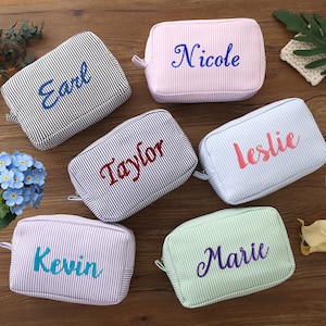 Könnte beinhalten: Eine Sammlung personalisierter Kosmetiktaschen in verschiedenen Pastellfarben, jede mit einem Namen in einer kontrastierenden Farbe bestickt. Die Taschen haben ein gestreiftes Muster und einen Reißverschluss. Namen sind Earl, Nicole, Taylor, Leslie, Kevin und Marie.