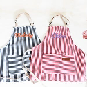 May include: Two striped aprons, one navy blue and the other red, with white vertical stripes. Each apron has a front pocket and the name "Melody" and "Chloe" embroidered on them. The aprons have cream-colored straps.