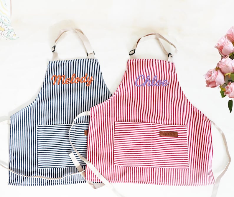 May include: Two striped aprons, one navy blue and the other red, with white vertical stripes. Each apron has a front pocket and the name "Melody" and "Chloe" embroidered on them. The aprons have cream-colored straps.