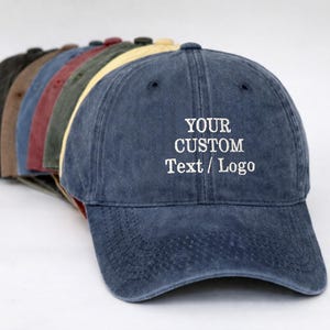 May include: A row of baseball caps in various colours including navy blue, yellow, green, red, brown, and black. The front-most navy blue cap features white embroidered text that reads YOUR CUSTOM Text / Logo, with a curved brim and six-panel design.