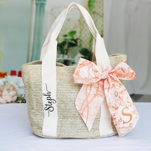 Op de afbeelding: Een beige strooien tas met witte banden en een gepersonaliseerde naam "Stepph" gedrukt op de band. De tas heeft een grote oranje en witte bloemenstrik.