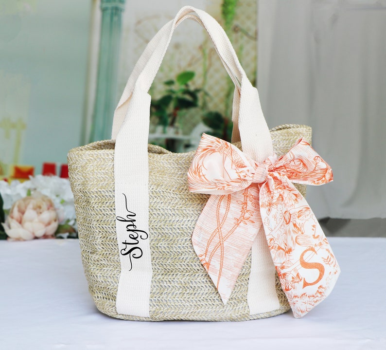 Op de afbeelding: Een beige strooien tas met witte banden en een gepersonaliseerde naam "Stepph" gedrukt op de band. De tas heeft een grote oranje en witte bloemenstrik.