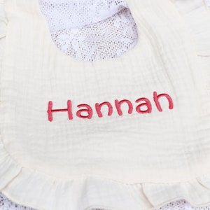 以下が含まれることがあります： 白い綿のスタイで、フリルが付いており、ピンク色の刺繍で「Hannah」と書いてあります。