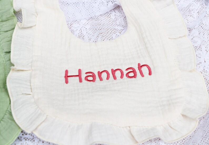 以下が含まれることがあります： 白い綿のスタイで、フリルが付いており、ピンク色の刺繍で「Hannah」と書いてあります。