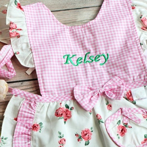 May include: Pink and white gingham and floral print apron with a bow and the name "Kelsey" embroidered on the front.