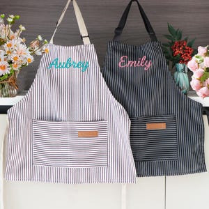 May include: Two striped aprons, one in black and white, the other in brown and white. The brown and white apron has the name "Aubrey" embroidered in teal. The black and white apron has the name "Emily" embroidered in pink. Each apron has a front pocket.