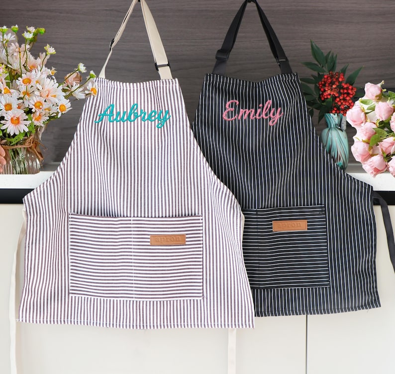 May include: Two striped aprons, one in black and white, the other in brown and white. The brown and white apron has the name "Aubrey" embroidered in teal. The black and white apron has the name "Emily" embroidered in pink. Each apron has a front pocket.