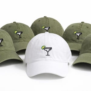 Pode incluir: Seis bonés de beisebol, um branco e cinco verde-oliva, são exibidos. Cada boné apresenta um bordado de um copo de margarita com uma fatia de limão. Os bonés são feitos de tecido macio.