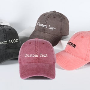 May include: Four baseball caps in different colors with customizable text. The caps are gray, black, pink, and red. The text on the caps reads "Custom Logo", "Custom Text", and "Custom".