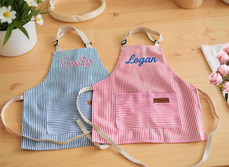 May include: Two striped aprons, one blue and one red, with the names "Emily" and "Logan" embroidered on them. Each apron has a pocket and cream-colored straps. The aprons are on a wooden surface.