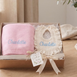 Può includere: Un set regalo per neonati in una scatola di cartone, con una coperta rosa e un bavaglino color crema, entrambi ricamati con il nome "Charlotte" in azzurro. Un'etichetta con la scritta "A Special Gift for Baby" è attaccata con un nastro.