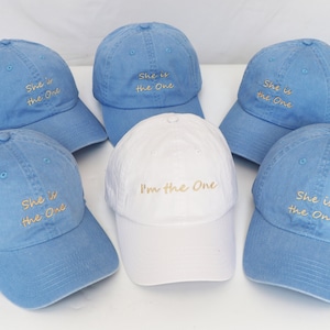 Puede incluir: Seis gorras de béisbol, cinco azules y una blanca, con texto bordado. Las gorras azules dicen "She is the One" y la gorra blanca dice "I'm the One".