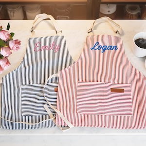 May include: Two striped aprons, one navy blue and one red, each with a pocket and personalized name embroidery. The navy apron has "Emily" embroidered in pink, and the red apron has "Logan" embroidered in blue. Both have cream-colored straps.
