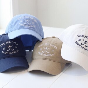 May include: Four baseball caps in navy blue, light blue, khaki, and white. Each cap displays the text "THE JEZ OPEN" with crossed golf clubs and the location "FRIPP ISLAND, SC".