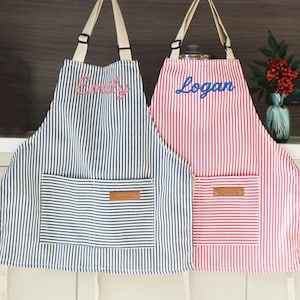 May include: Two striped aprons, one blue and white, the other red and white, hang side by side. The blue apron has "Emily" embroidered in pink, and the red apron has "Logan" in blue. Each has a front pocket.