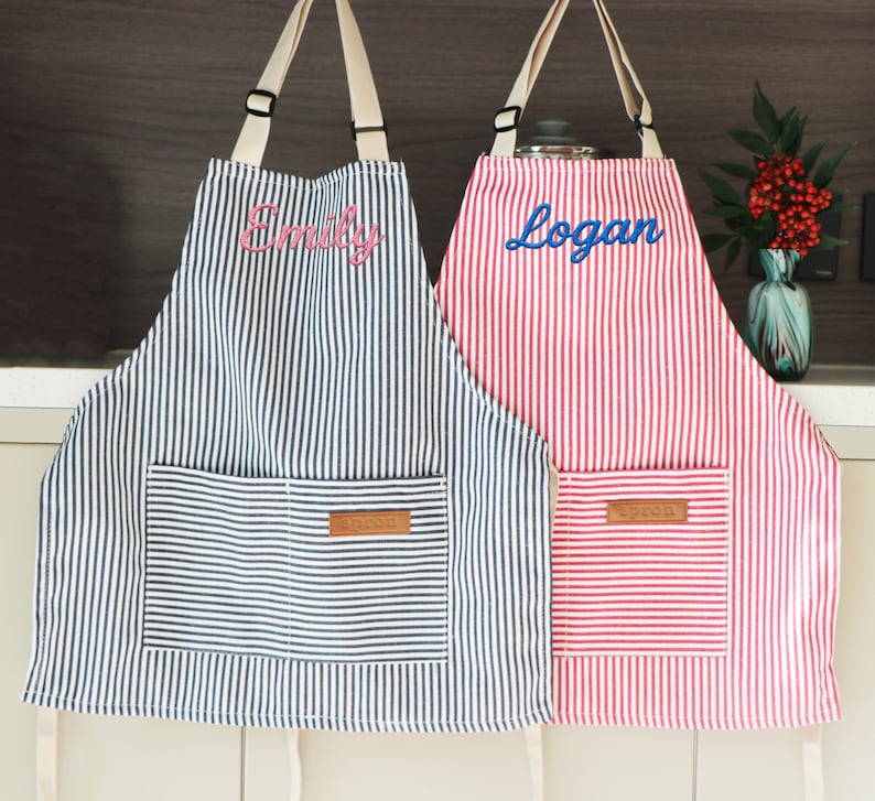 May include: Two striped aprons, one blue and white, the other red and white, hang side by side. The blue apron has "Emily" embroidered in pink, and the red apron has "Logan" in blue. Each has a front pocket.
