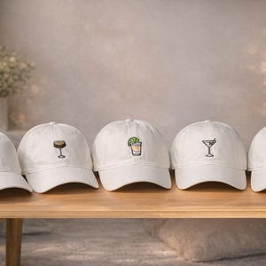 Puede incluir: Cinco gorras de béisbol blancas con diseños de cócteles bordados. Cada gorra presenta una bebida diferente: un martini, una margarita, una botella de champán, un cóctel y una bebida con una guarnición. Las gorras se exhiben sobre una mesa de madera.