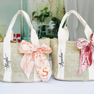 Op de afbeelding: Twee beige strooien tassen met witte handvatten en gepersonaliseerde namen. De tas links heeft een perzikkleurig lint met een bloemenpatroon en de naam "Steph" erop geschreven. De tas rechts heeft een rood lint met een bloemenpatroon en de naam "Anusha" erop geschreven.