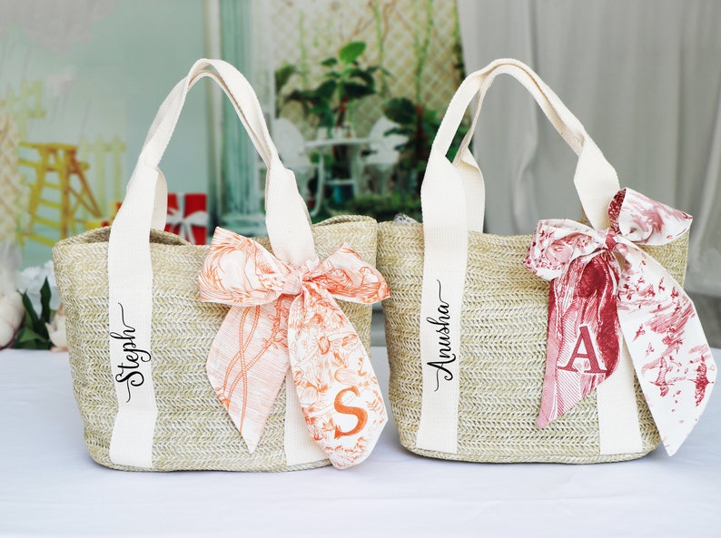 Op de afbeelding: Twee beige strooien tassen met witte handvatten en gepersonaliseerde namen. De tas links heeft een perzikkleurig lint met een bloemenpatroon en de naam "Steph" erop geschreven. De tas rechts heeft een rood lint met een bloemenpatroon en de naam "Anusha" erop geschreven.
