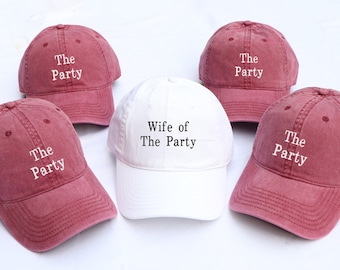May include: Five baseball caps, four are maroon and one is white. The maroon caps have white text that says "The Party". The white cap has black text that says "Wife of The Party".