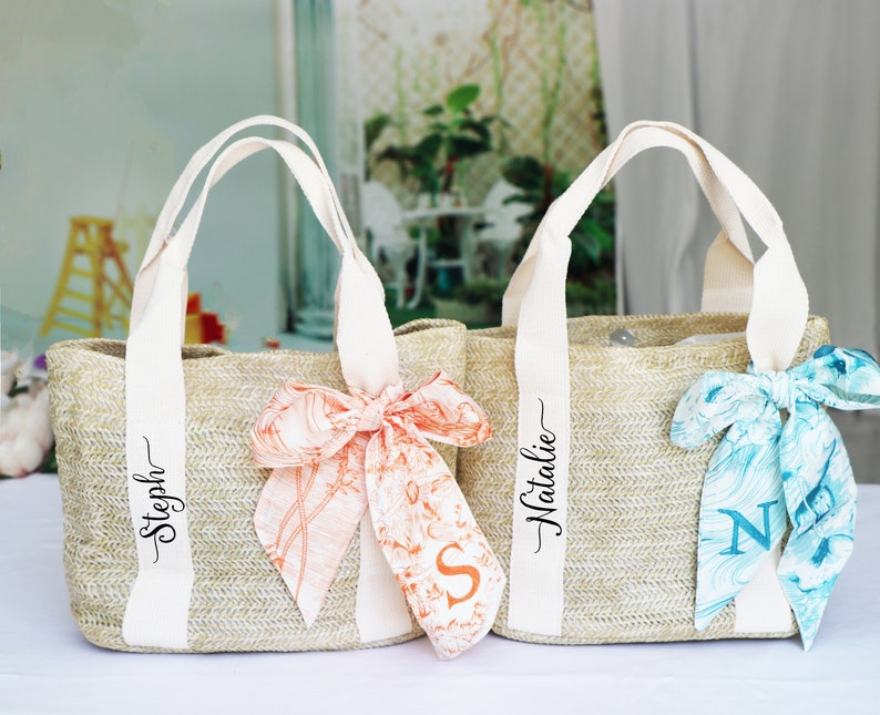 Op de afbeelding: Twee beige strooien tassen met witte handvatten en gepersonaliseerde namen. De tas links heeft een perzikkleurig en wit gepatroneerd lint om de handvatten gebonden en de naam "Steph" op de tas. De tas rechts heeft een blauw en wit gepatroneerd lint om de handvatten gebonden en de naam "Natalie" op de tas.