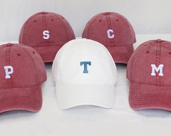 May include: Five baseball caps, four are red and one is white. The red caps have white letters on them, spelling out "P", "S", "C", and "M". The white cap has a teal blue letter "T" on it.