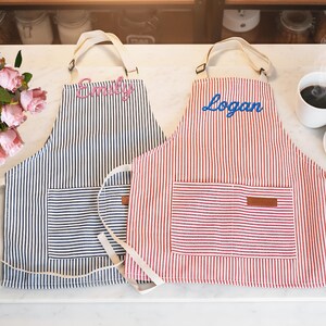 May include: Two striped aprons, one navy and white, the other red and white, each with a front pocket and a name embroidered on the bib. The navy apron has "Emily" embroidered in pink, and the red apron has "Logan" embroidered in blue.