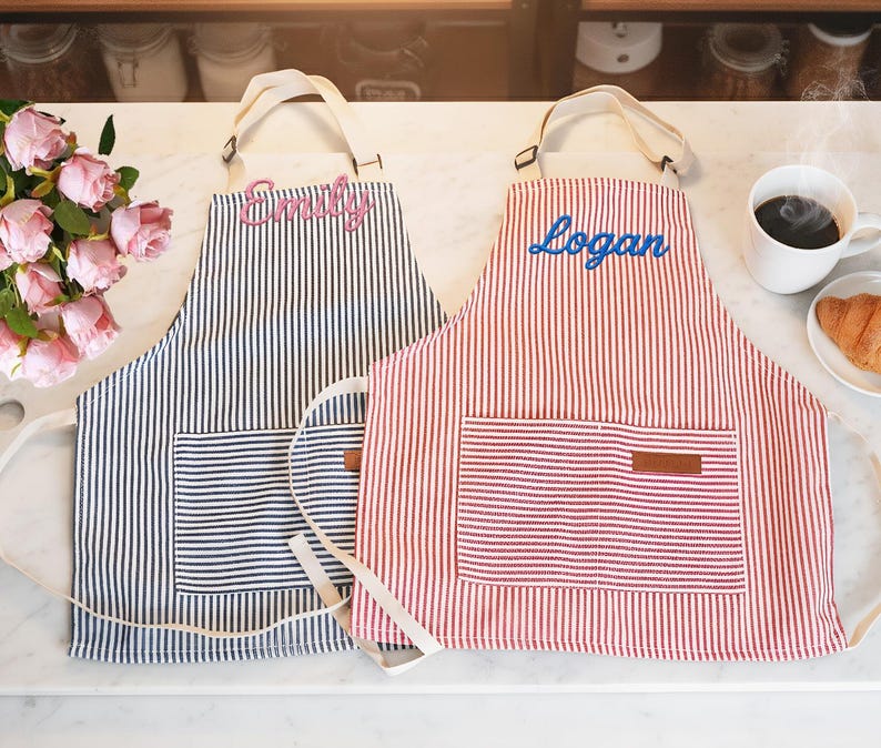 May include: Two striped aprons, one navy and white, the other red and white, each with a front pocket and a name embroidered on the bib. The navy apron has "Emily" embroidered in pink, and the red apron has "Logan" embroidered in blue.