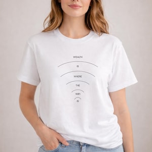 May include: White t-shirt with the text "WEALTH IS WHERE THE WIFI IS" in black. The text is arranged in a descending arc design. The model is wearing blue jeans.