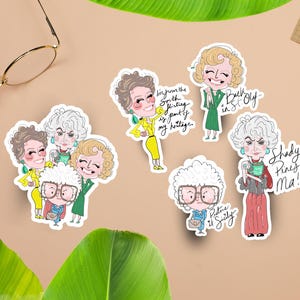 May include: A collection of colorful stickers featuring cartoon illustrations of characters with text. The stickers depict women in various outfits, with phrases like "Picture it Sicily" and "Shady Pines Ma!" on them. The stickers are on a tan surface.