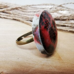 Könnte beinhalten: Ein verstellbarer Ring mit einem großen, ovalen Stein. Der Stein hat ein marmoriertes Design in Rot, Schwarz und Weiß. Das Ringband ist bronzefarben und der Stein liegt auf einer Holzoberfläche.