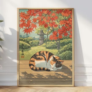 May include: A framed print of a sleeping calico cat in a Japanese garden. The artwork features a cat curled up on a wooden deck, with a backdrop of red maple leaves, a stone lantern, and lush greenery. The print has a vintage aesthetic.
