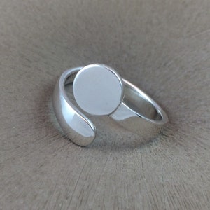 May include: A silver ring with a round, blank, silver disc on top. The ring is open and can be adjusted to fit different finger sizes.